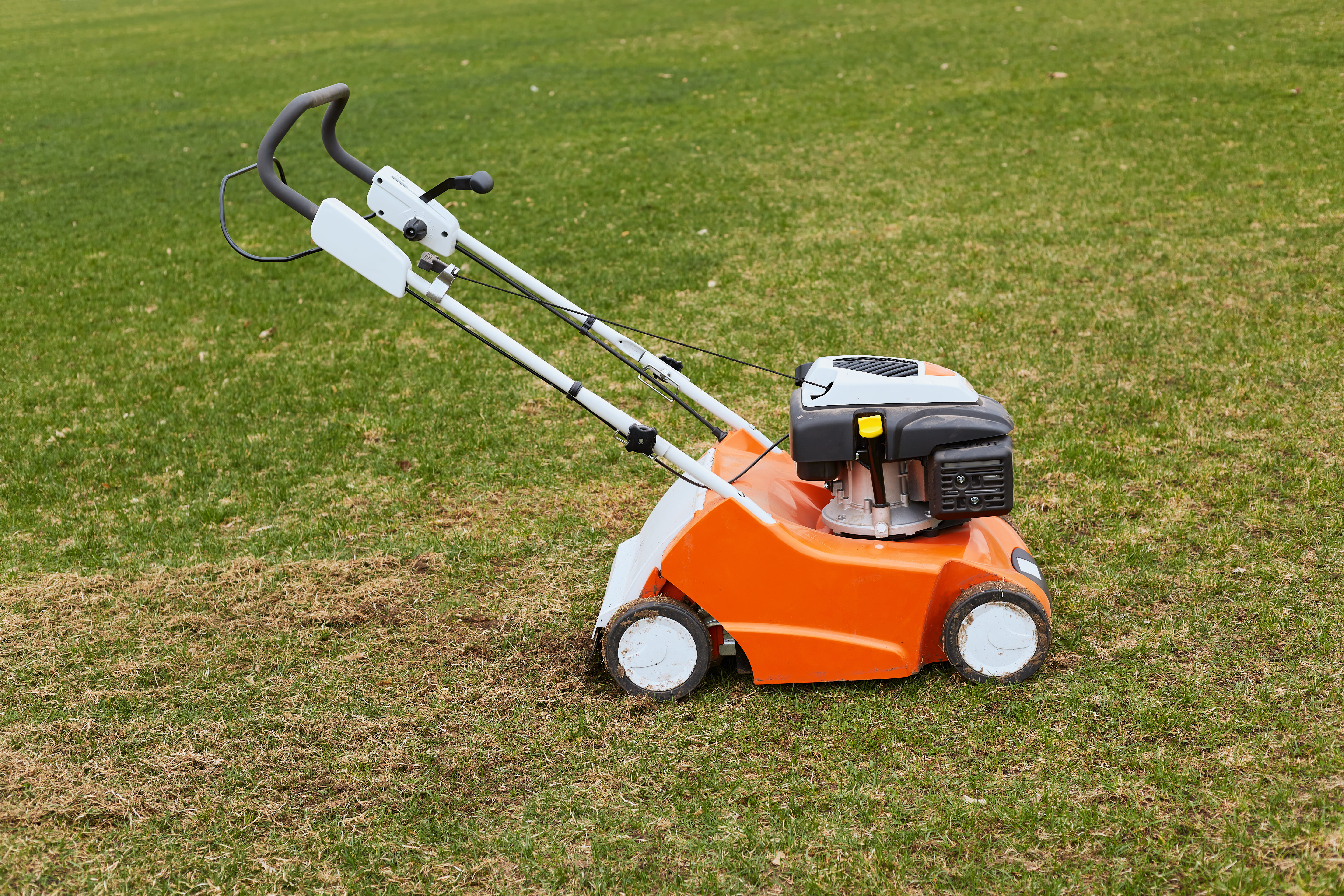A photograph of a person, likely a gardener or landscaper, standing on a lawn with a green grass background, holding an orange-colored grass cutter or lawn mower, possibly in a residential or commercial outdoor setting